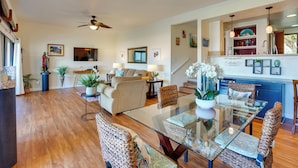 A spacious living area with a glass dining table, comfortable seating, and a kitchen area with blue cabinets and hanging lights.