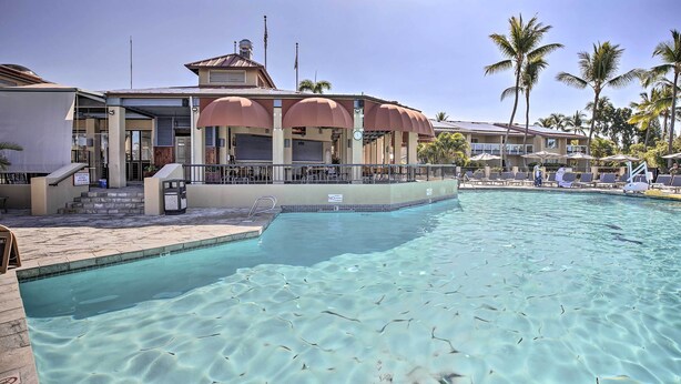 A pool area with clear water, palm trees, and a building with a covered seating area.