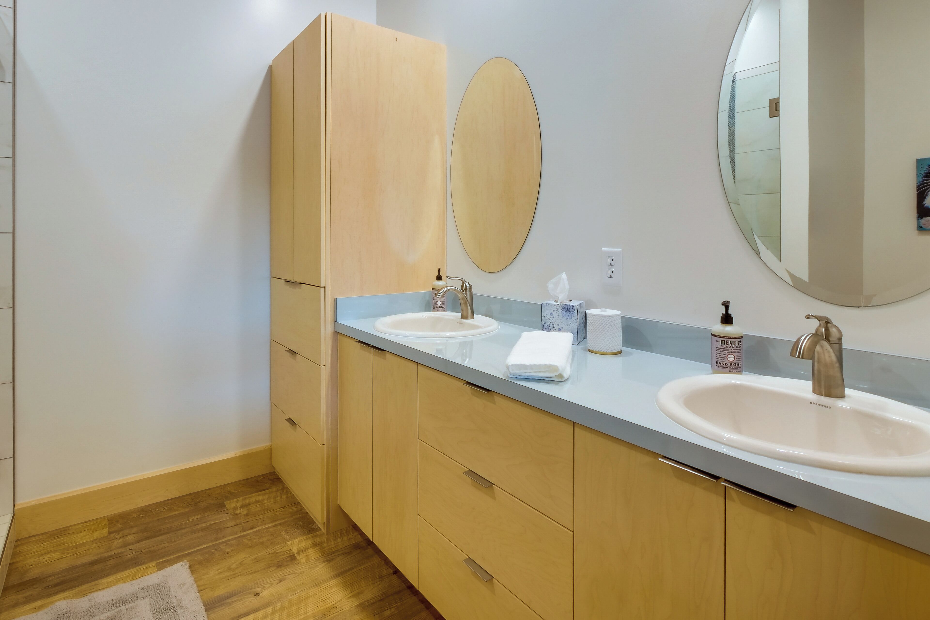A modern bathroom with two sinks, wooden cabinets, a large mirror, and a light-colored countertop.