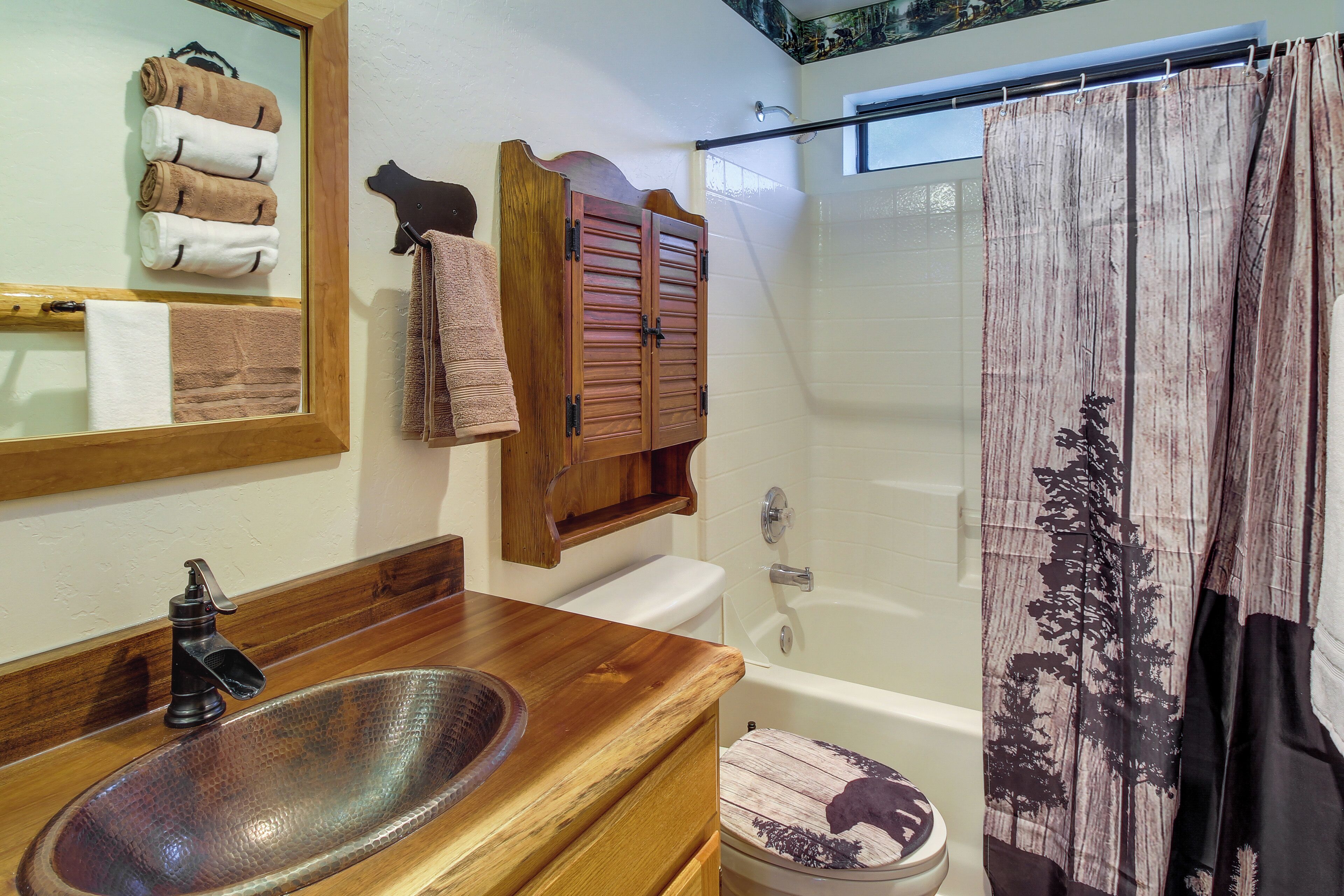 A bathroom with a wooden vanity, a glass sink, a white toilet, and a shower curtain with a forest design.