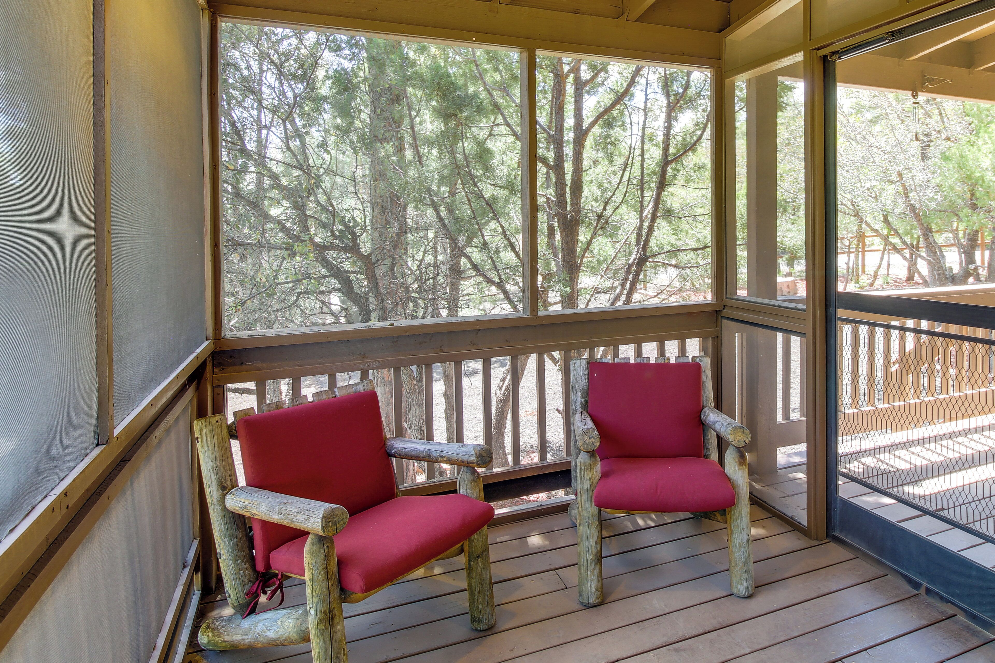 A wooden deck with two red cushioned chairs and a view of trees through a screened window.