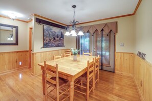 Een eetruimte met een houten tafel en stoelen, een kroonluchter, een schilderij aan de muur en een raam met jaloezieën.
