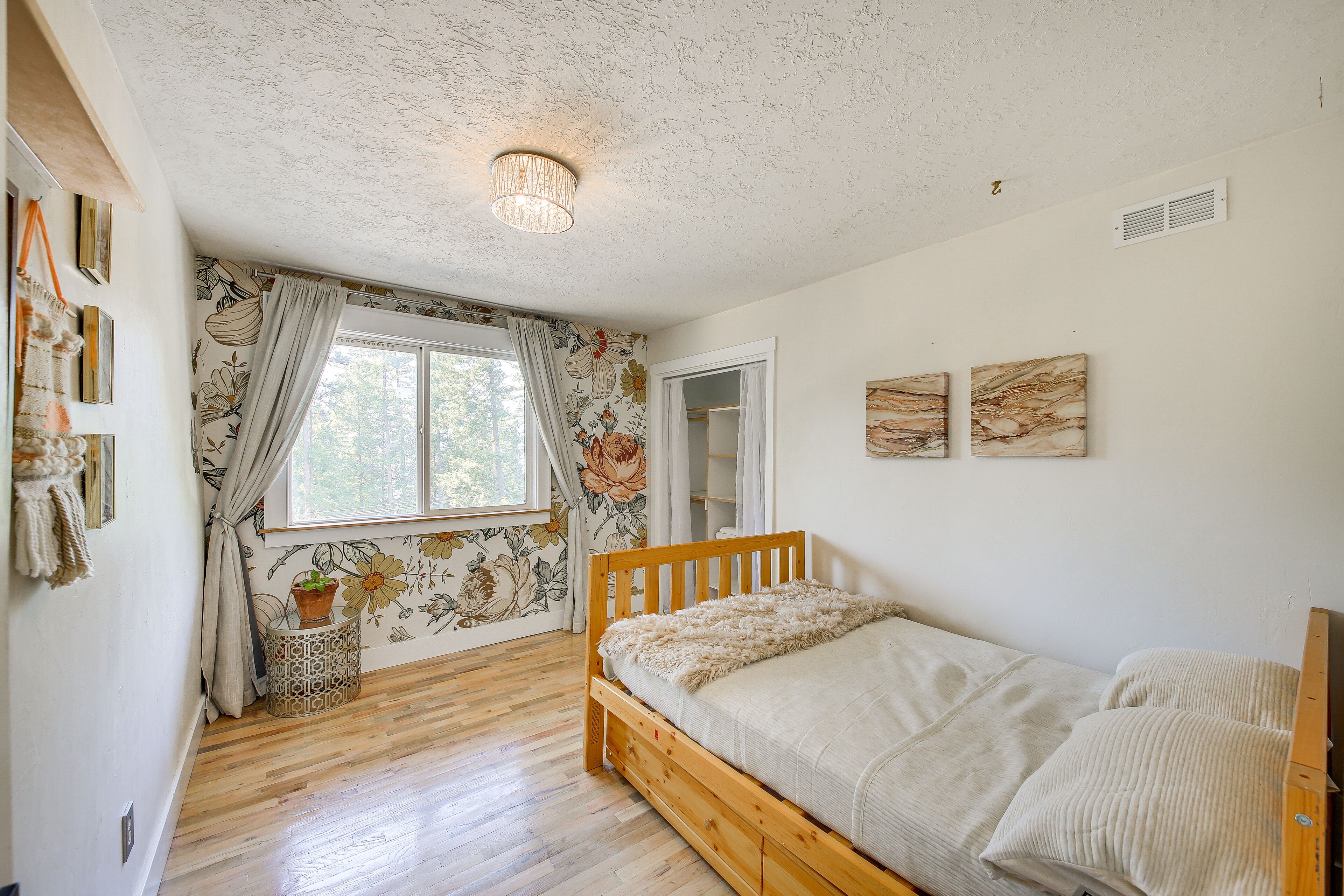 A bedroom with a wooden bed, a window with floral curtains, a small side table with a plant, and a ceiling light fixture.