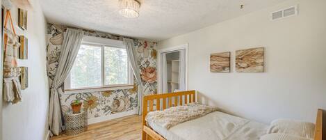 A bedroom with a wooden bed, a window with floral curtains, a small side table with a plant, and a ceiling light fixture.