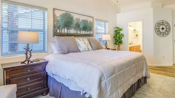 Quarto com uma cama grande, duas mesas de cabeceira, cômoda, janela com persianas, quadro, relógio e planta.