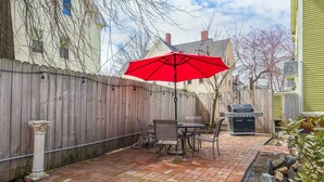 Une terrasse à l’arrière de la maison, avec un sol en briques, un parasol rouge, un barbecue et une clôture en bois.