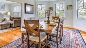 Un coin repas avec une table en bois, des chaises et un tapis à motifs. On y trouve des fenêtres avec des stores, un ventilateur de plafond et un radiateur.