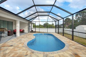 A covered outdoor swimming pool with a stone deck, a grill, and a patio area with seating.