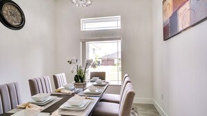 A dining room with a long table set for six, a clock on the wall, and a window providing a view of the outside. - Furnished Lanai & Yard: Comfy Lehigh Acres Getaway (Lehigh Acres)