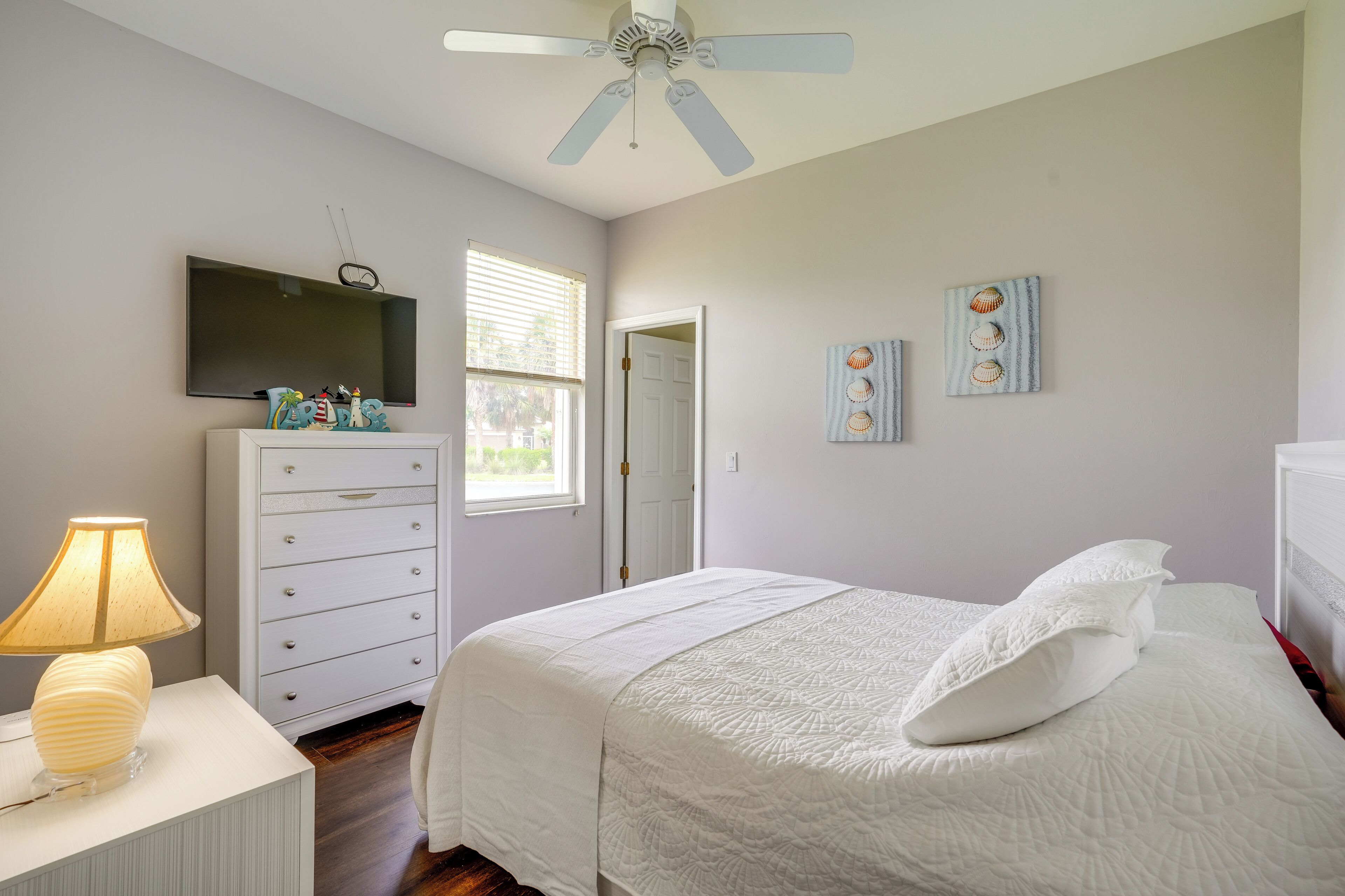 A bedroom with a bed, a dresser, a lamp, a ceiling fan, a TV, and a window.