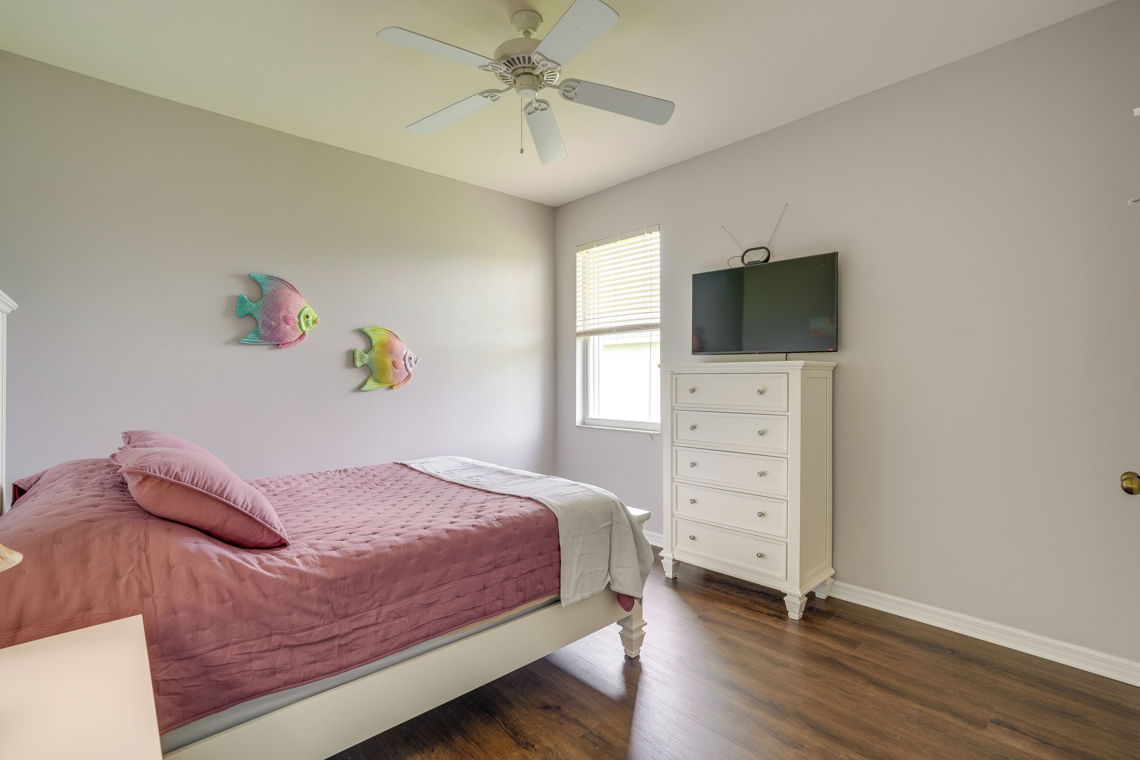 A bedroom with a bed, dresser, ceiling fan, and a TV on the wall.