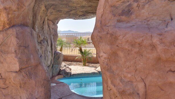 Un espace piscine agrémenté d’une arche naturelle en roche, d’un petit palmier et offrant une vue lointaine sur un bâtiment.