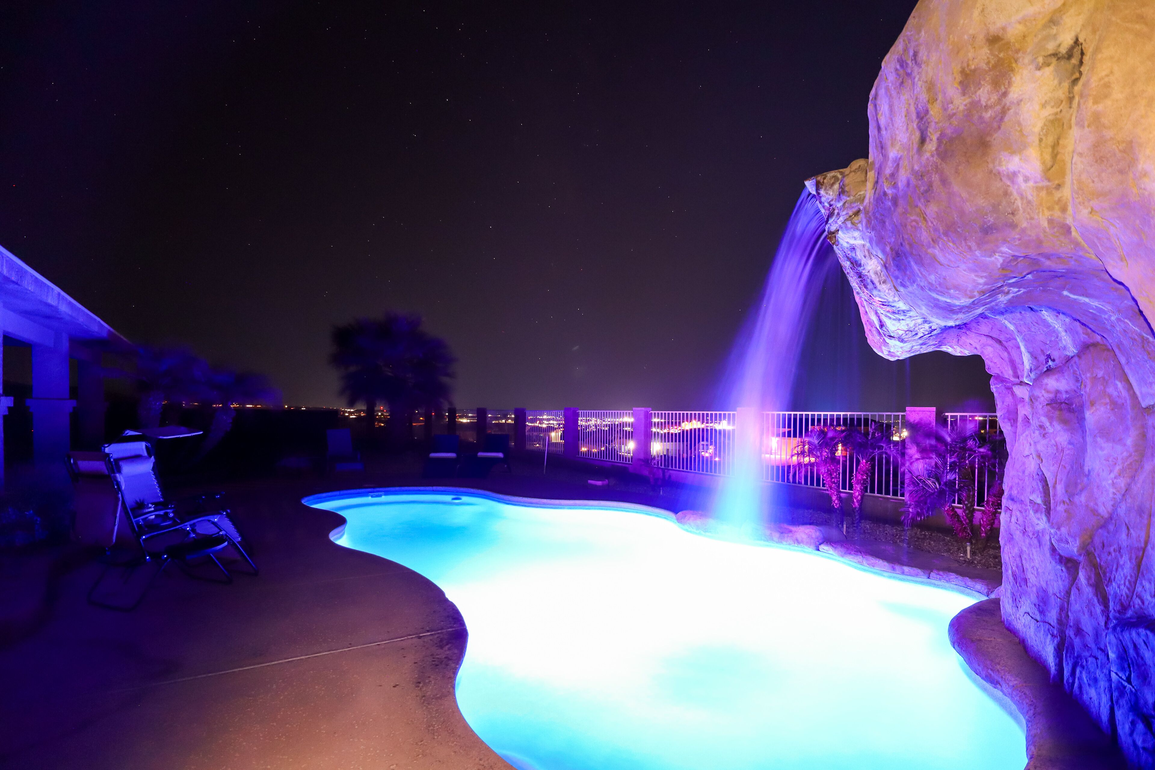 Ein Nacht-Schwimmbad mit Wasserfall, umgeben von Liegestühlen und Blick auf eine Stadtlandschaft.