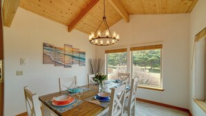 Un coin repas avec une table en bois, des chaises blanches et un lustre. Des tableaux ornent le mur, et une fenêtre offre une vue sur les arbres.