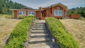 Une maison en bois à deux étages, dotée d’un escalier en pierre menant à la porte d’entrée, entourée d’une végétation luxuriante et bordée par une forêt.