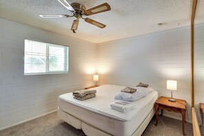 Une chambre avec un mur en briques blanches, un lit simple avec un matelas et des oreillers blancs, une table de chevet en bois avec une lampe, un ventilateur de plafond et une fenêtre avec des stores.