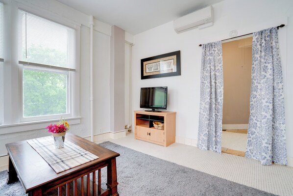Une pièce avec une table en bois, un téléviseur posé sur un meuble en bois, un tableau encadré au mur et une fenêtre avec des stores.