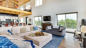 A spacious living area with a large sofa, a coffee table, and a television. There is a kitchen area with cabinets and a dining table. The room has large windows and a view of the outdoors. - Colorado Retreat w/ Gas Grill & Mountain Views! (Fairplay)
