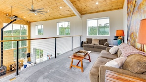 A spacious living area with a high ceiling, wooden beams, a balcony with a railing, and a large window offering a view of the outdoors. - Colorado Retreat w/ Gas Grill & Mountain Views! (Fairplay)