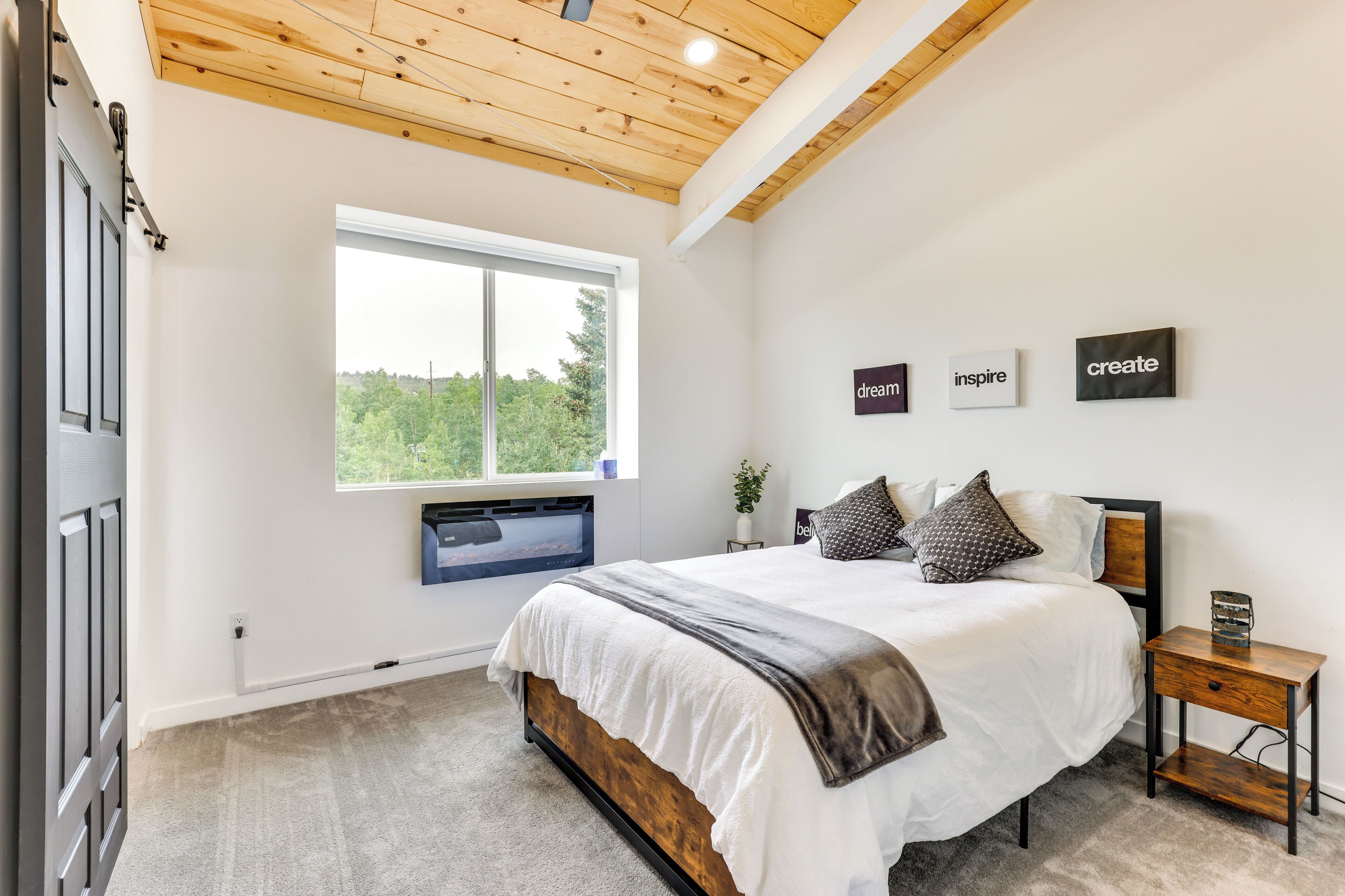 A bedroom with a wooden ceiling, a large bed with a white and brown blanket, a bedside table, and a window with a view of greenery.