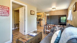 A living room with a sofa, coffee table, and television. There is a kitchen in the background with wooden cabinets and a dining table. - On-site Creek & Fishing: Cozy Coos Bay Retreat! (Coos Bay)