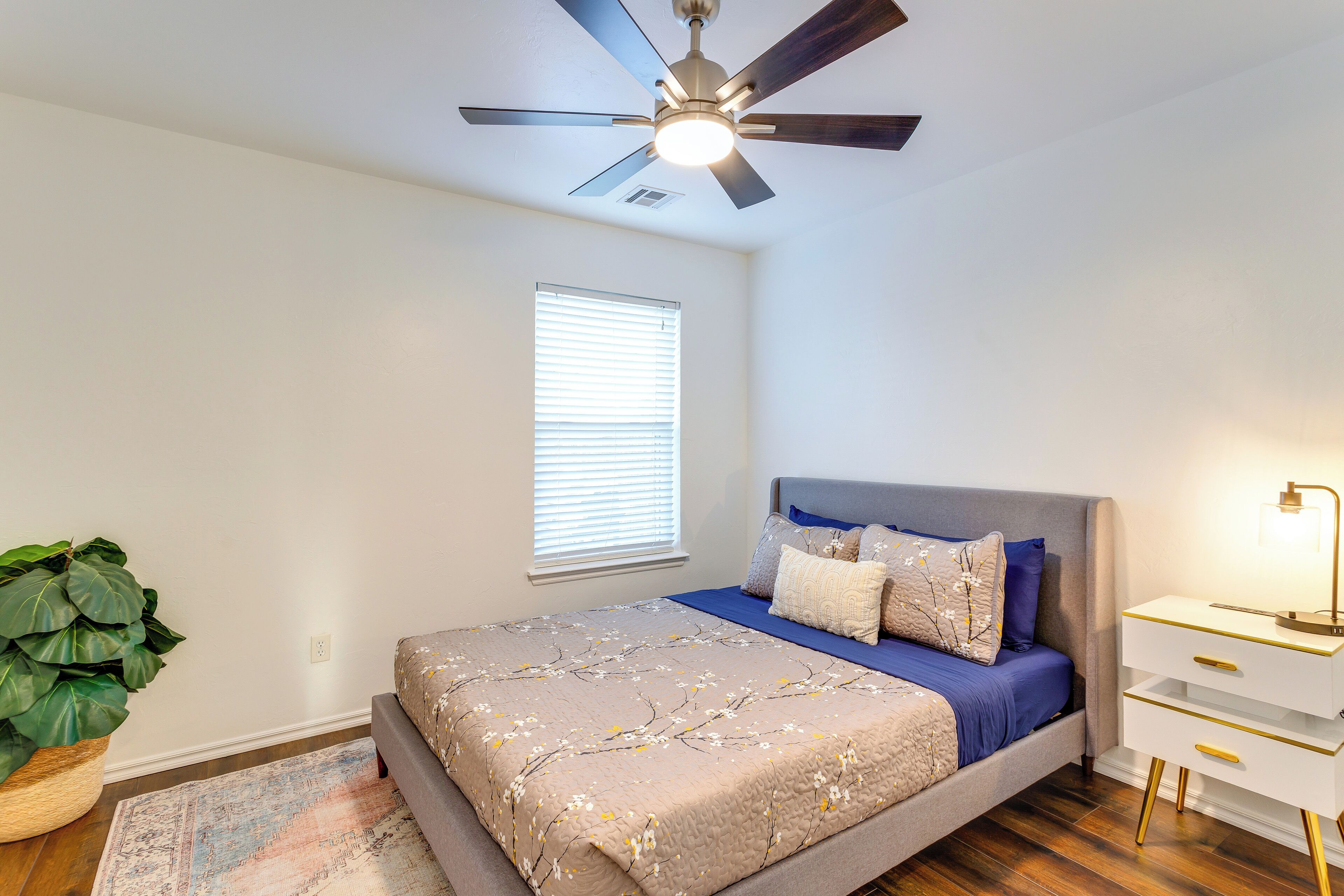 A bedroom with a bed, a nightstand, a ceiling fan, a window with blinds, and a potted plant.
