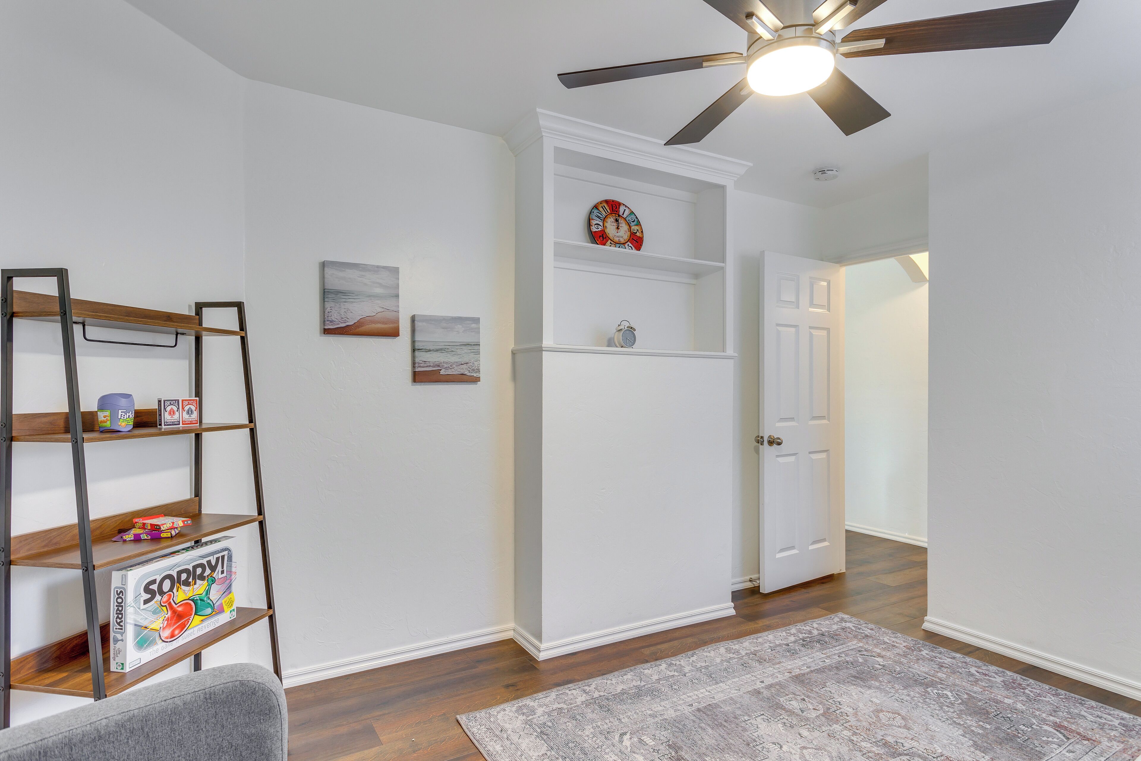 A room with a ceiling fan, a white door, a shelf with a clock, and a rug.