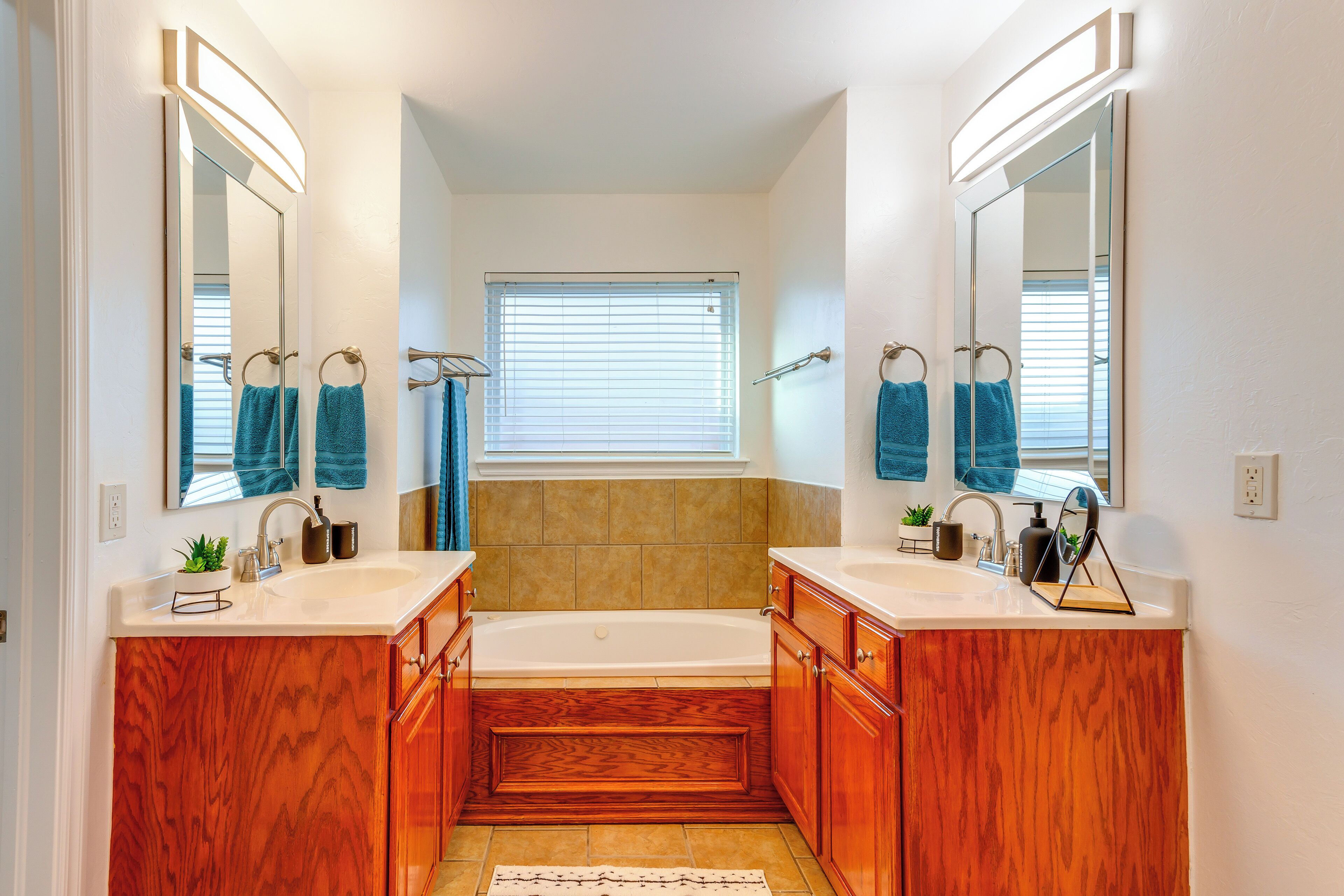 Un baño con dos lavamanos, una bañera, armarios de madera y una ventana con persianas.