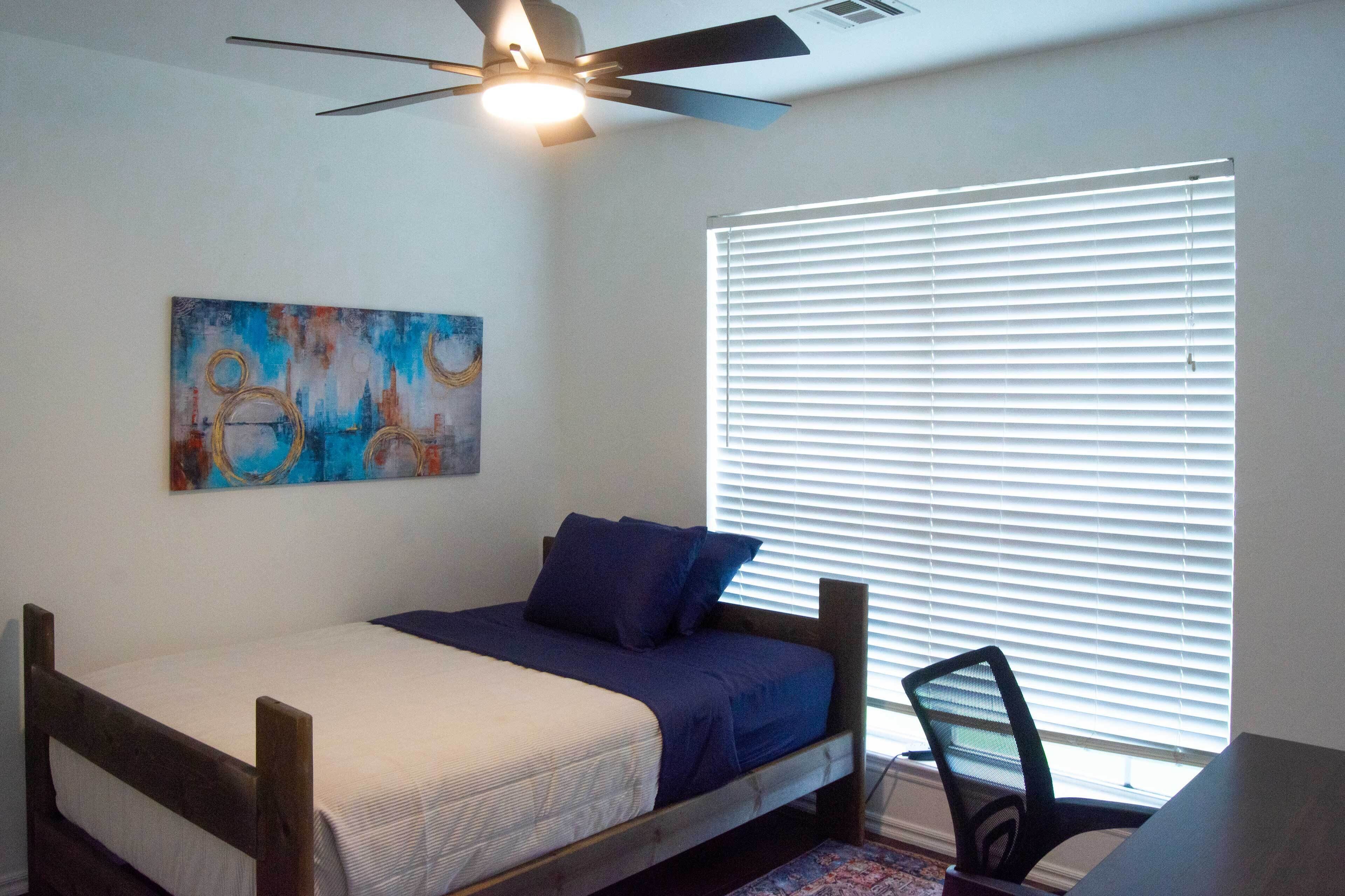 Une chambre à coucher avec un lit, un ventilateur de plafond, une fenêtre avec des stores et un tableau au mur.
