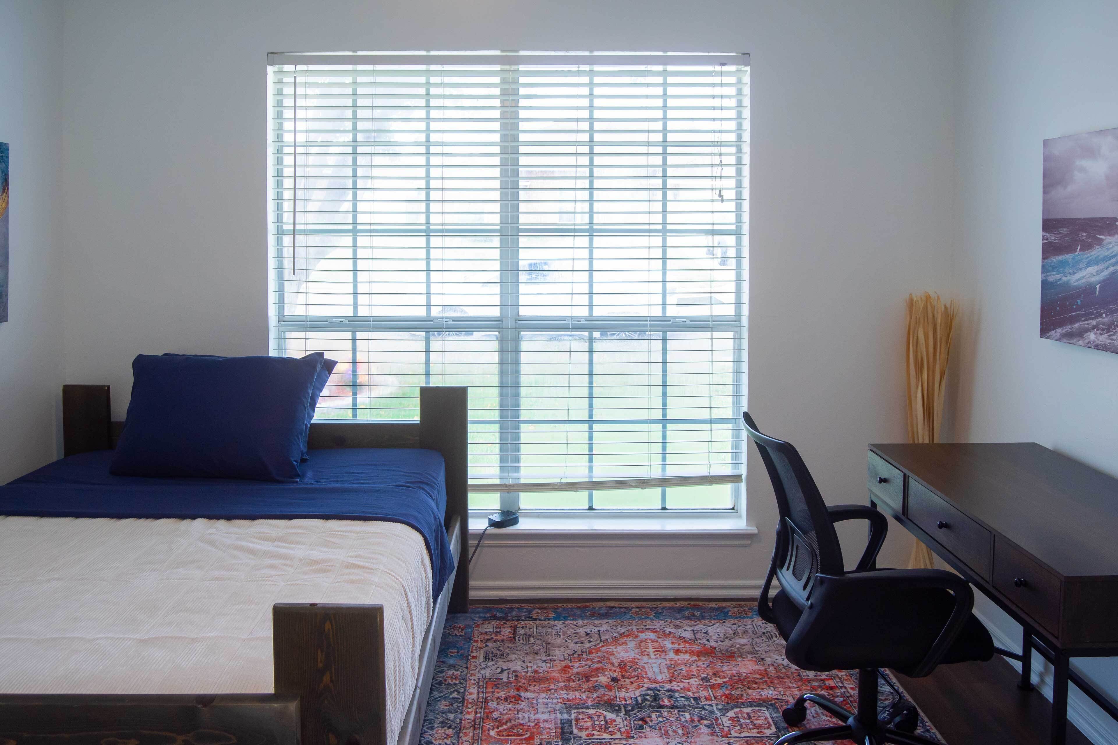 A bedroom with a bed, a desk, and a window with blinds.
