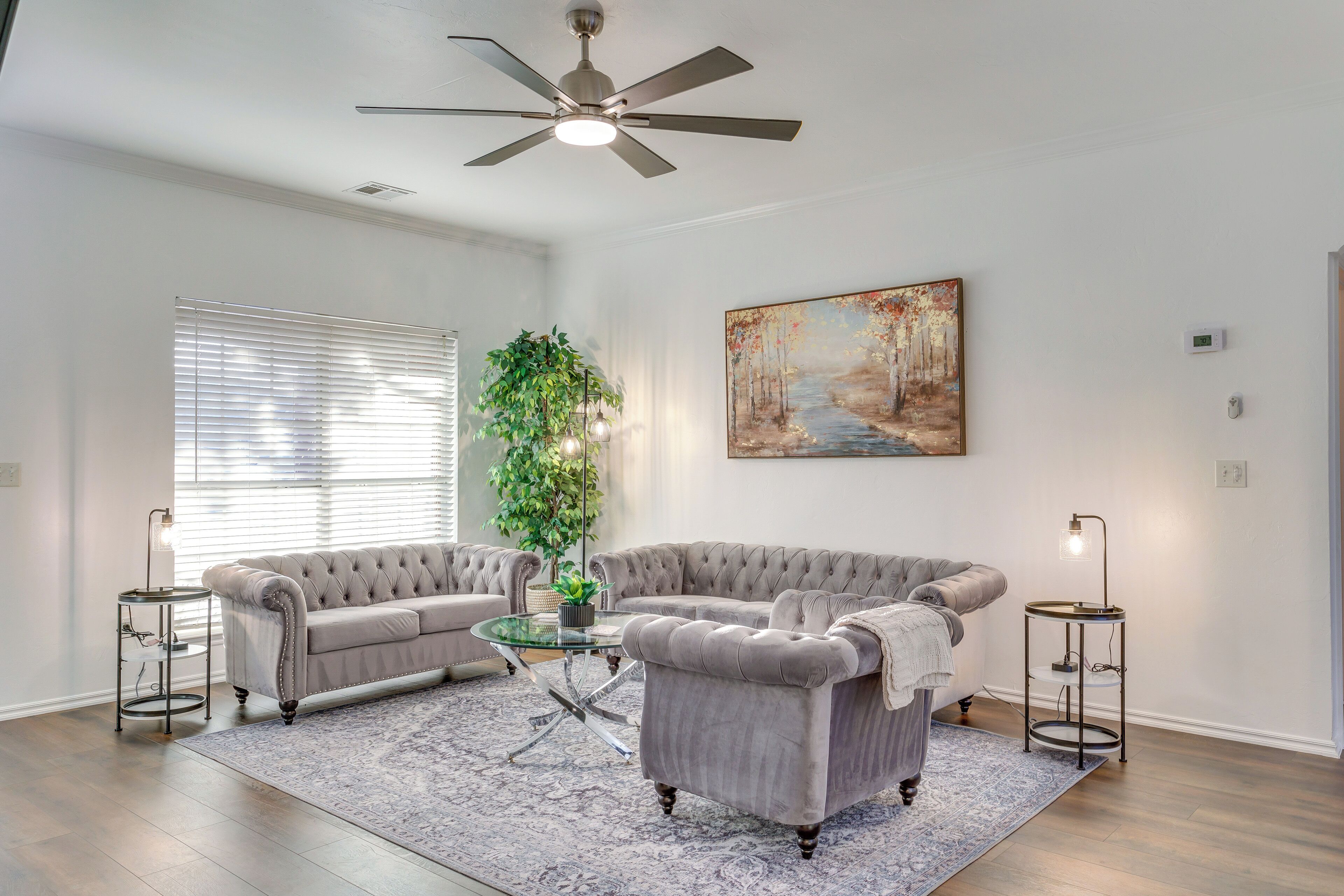 A living room with two grey upholstered sofas, a glass coffee table, a ceiling fan, a painting, a potted plant, and a side table with a lamp.