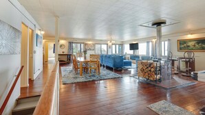 A spacious living room with a wooden floor, a blue sofa, a dining table with chairs, and a fireplace. - Waterfront Coos Bay Retreat w/ Sauna! (Coos Bay)