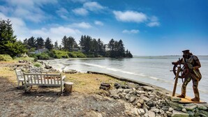 A wooden bench and a statue by a rocky shoreline with a view of a calm body of water and trees in the background. - Waterfront Coos Bay Retreat w/ Sauna! (Coos Bay)