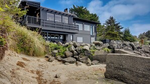 A modern two-story house with a balcony, situated on a rocky shoreline with trees in the background. - Waterfront Coos Bay Retreat w/ Sauna! (Coos Bay)