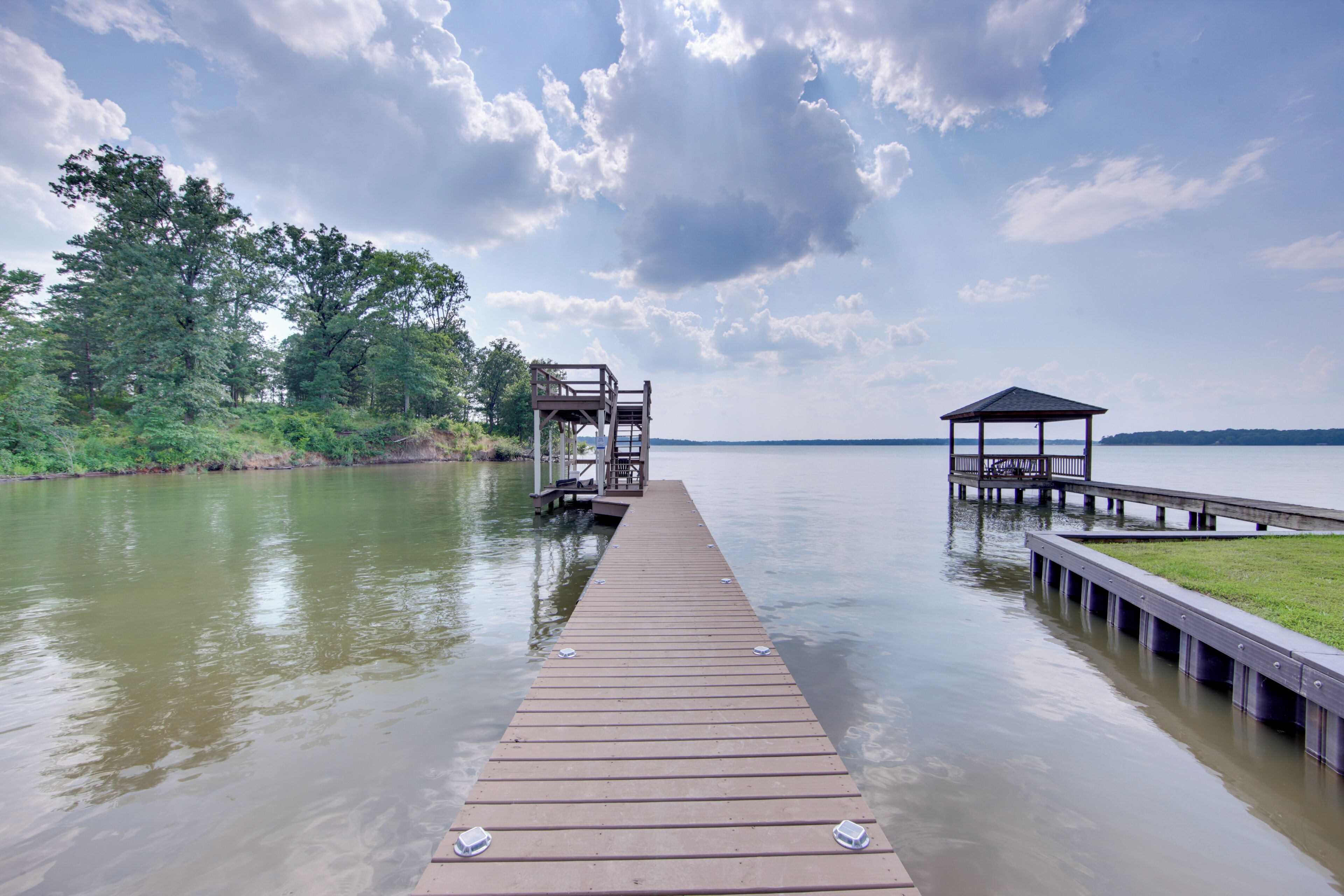 A wooden pier extending into a calm lake with a gazebo and a wooden structure on the left.