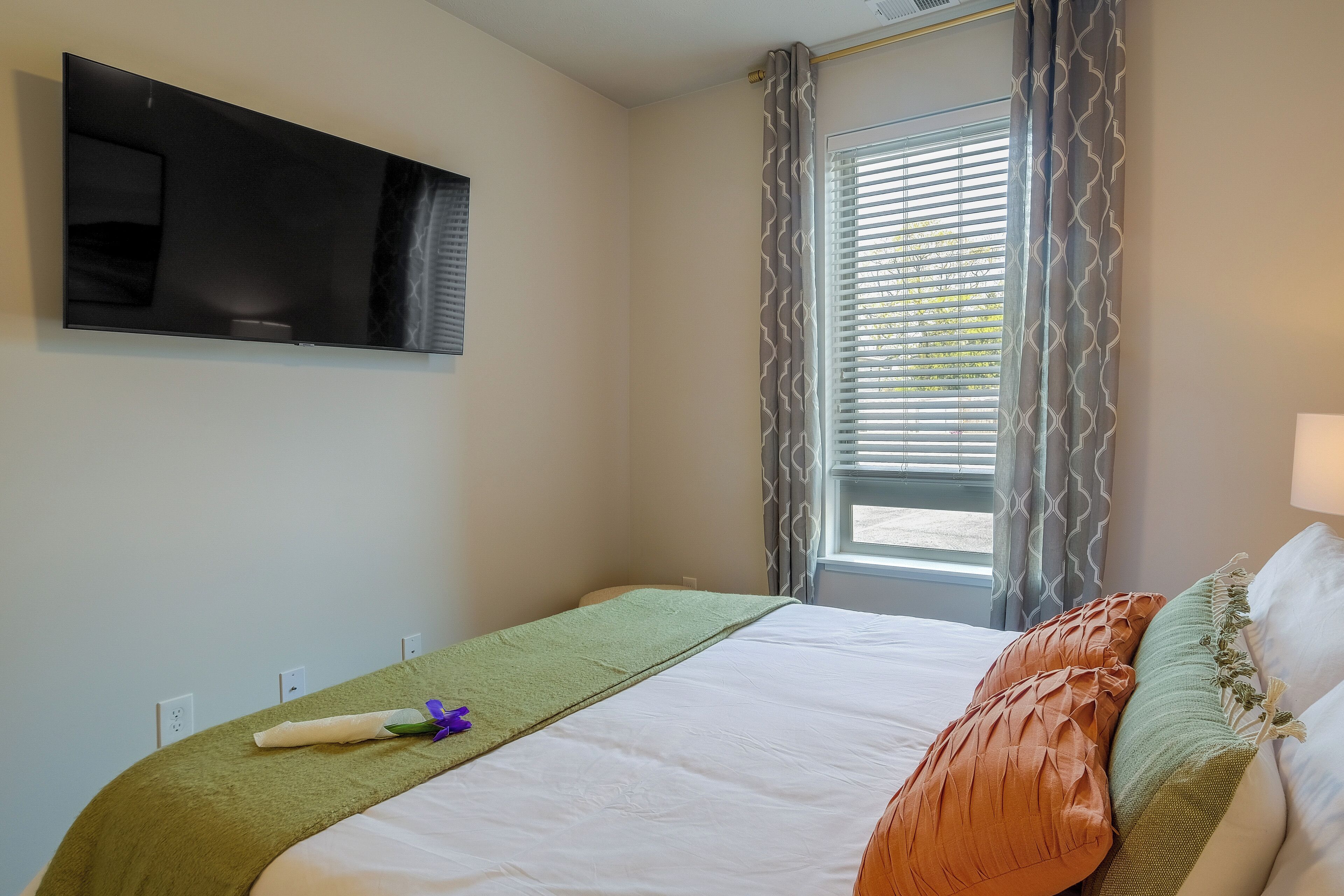 A neatly made bed with a green blanket and decorative pillows, a flat-screen TV mounted on the wall, and a window with patterned curtains.