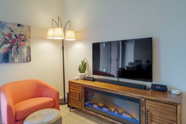 A living room with a flat-screen TV, a fireplace with a glass front, a sofa, a floor lamp, and a painting on the wall. - Traverse City Vacation Rental: 1 Mi to Beach! (Traverse City)