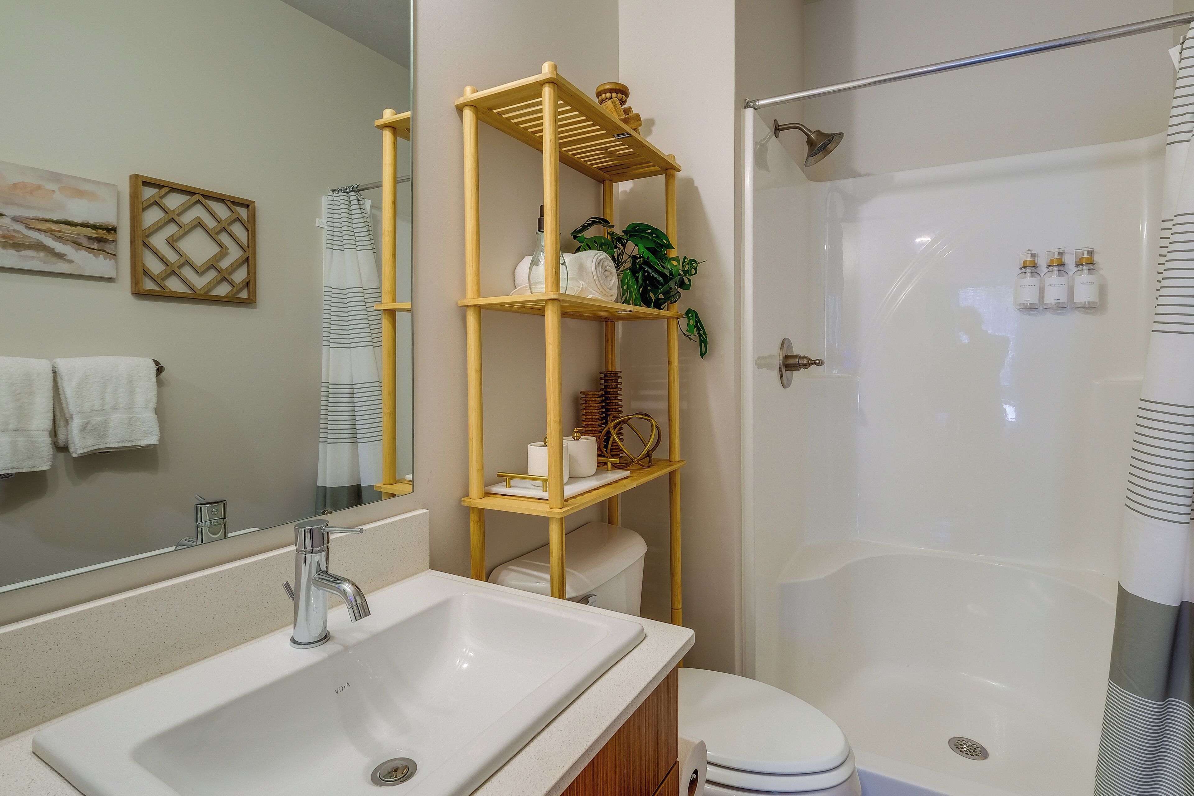 A bathroom with a large sink, toilet, and shower. There is a wooden shelf with toiletries and a plant.