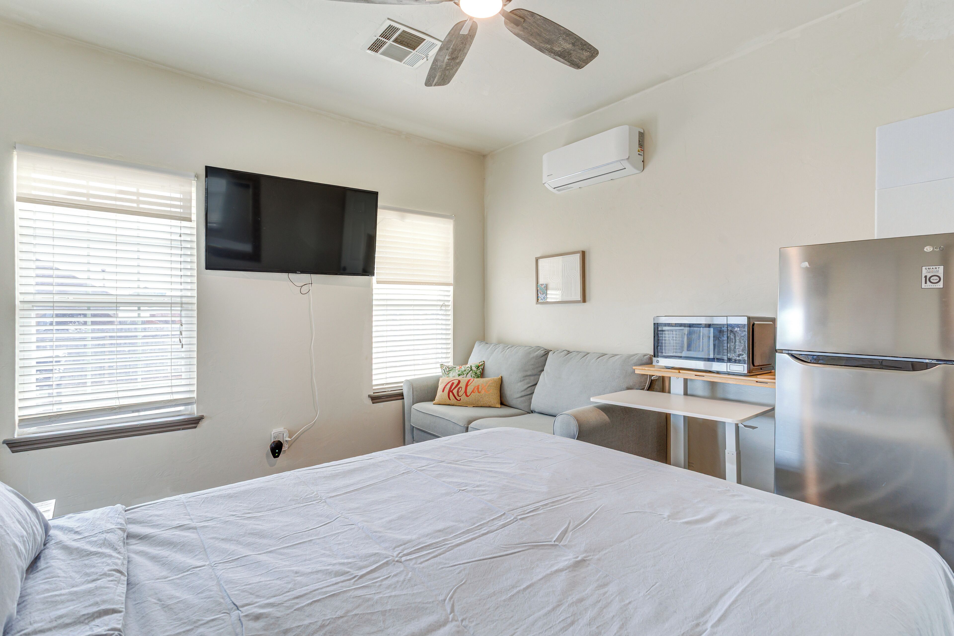 Un dormitorio con una cama, un escritorio con microondas, refrigerador, ventilador de techo y televisor montado en la pared.