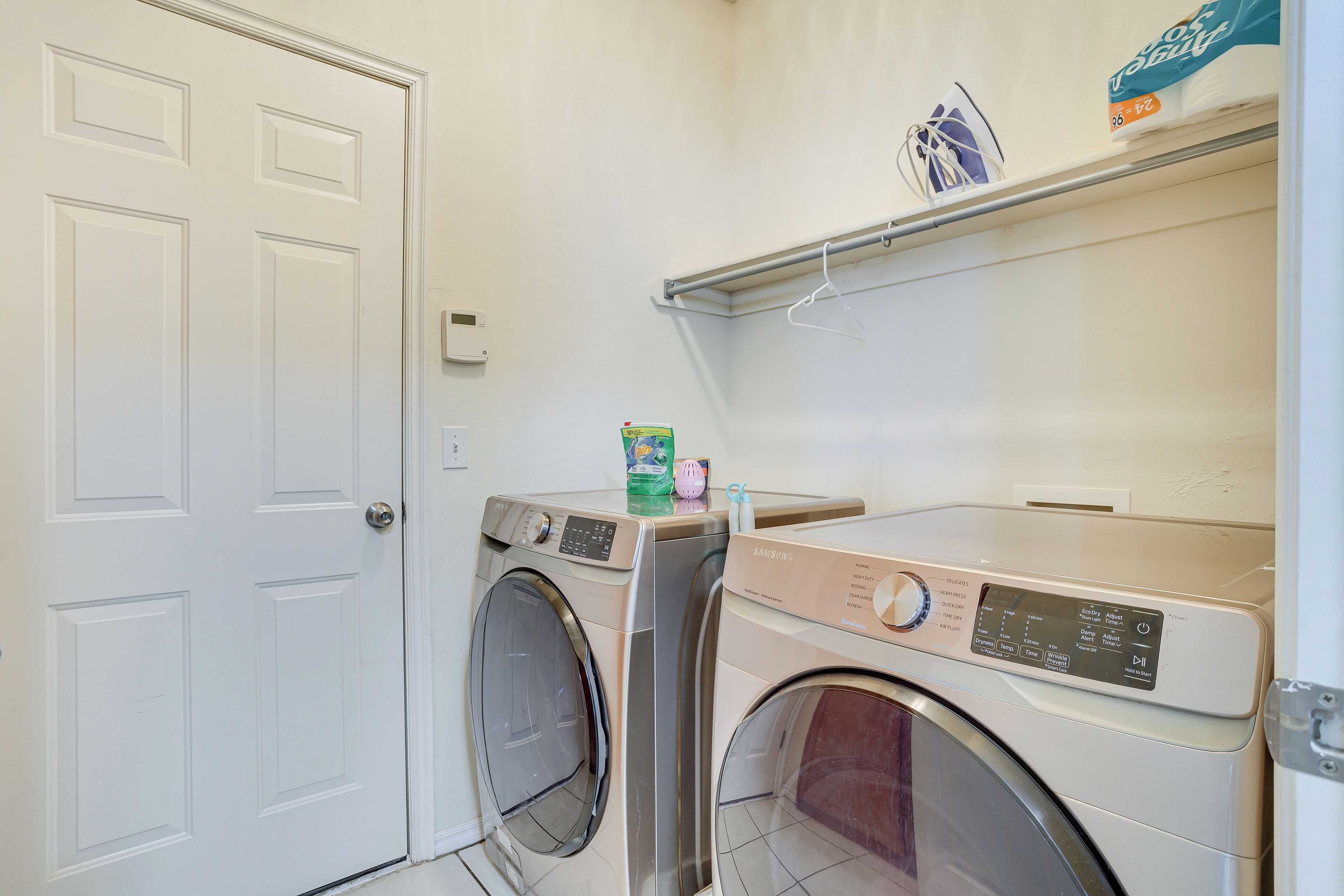 A laundry room with two washing machines, a dryer, and a shelf with laundry supplies.