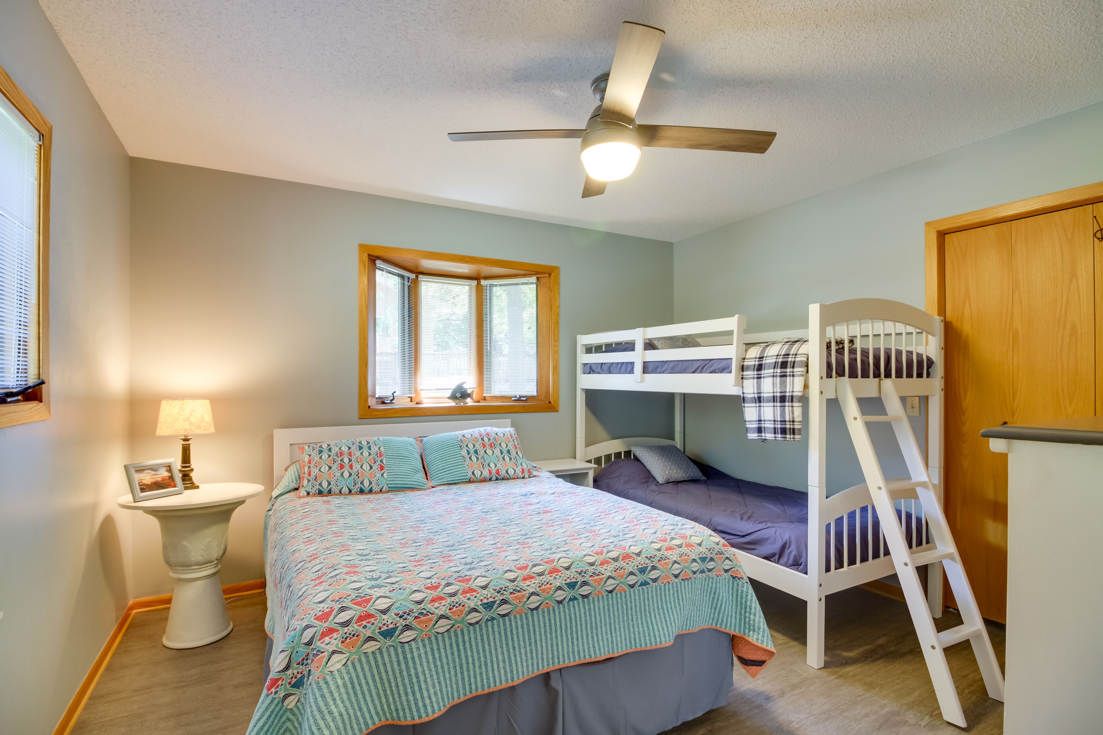 A bedroom with a bunk bed, a ceiling fan, a bedside table with a lamp, and a window with blinds.