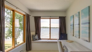 A room with a large window overlooking a body of water, a bed with patterned bedding, a desk with a notebook, and a wall with three paintings.