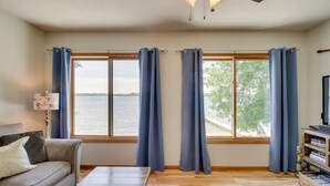 A living room with a sofa, a coffee table, a TV, and a bookshelf. There are three windows with curtains, and a ceiling fan.