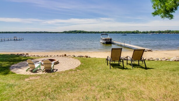 A lakeside view with a dock, chairs, and a fire pit.