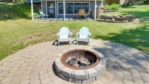 A fire pit with two Adirondack chairs, a patio area, and a house with a covered deck in the background. - Boat Dock & Private Beach: Lake Mary Retreat! (Alexandria)