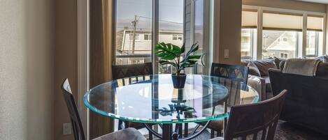 A glass-top dining table with four chairs, a potted plant, and a view of buildings through a sliding glass door.