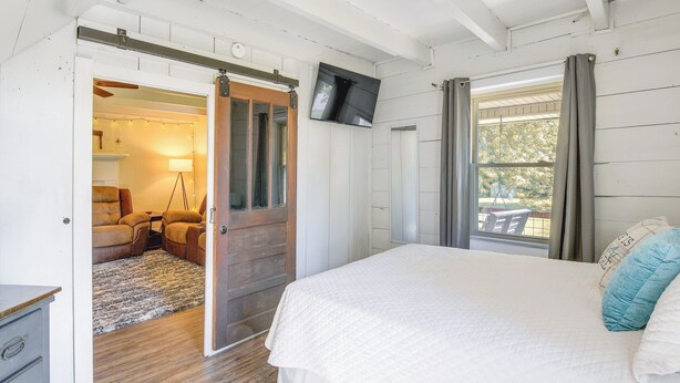 Ein Schlafzimmer mit Bett, einem an der Wand montierten Fernseher und einer Schiebetür zu einem Wohnbereich mit Sofa und Lampe.