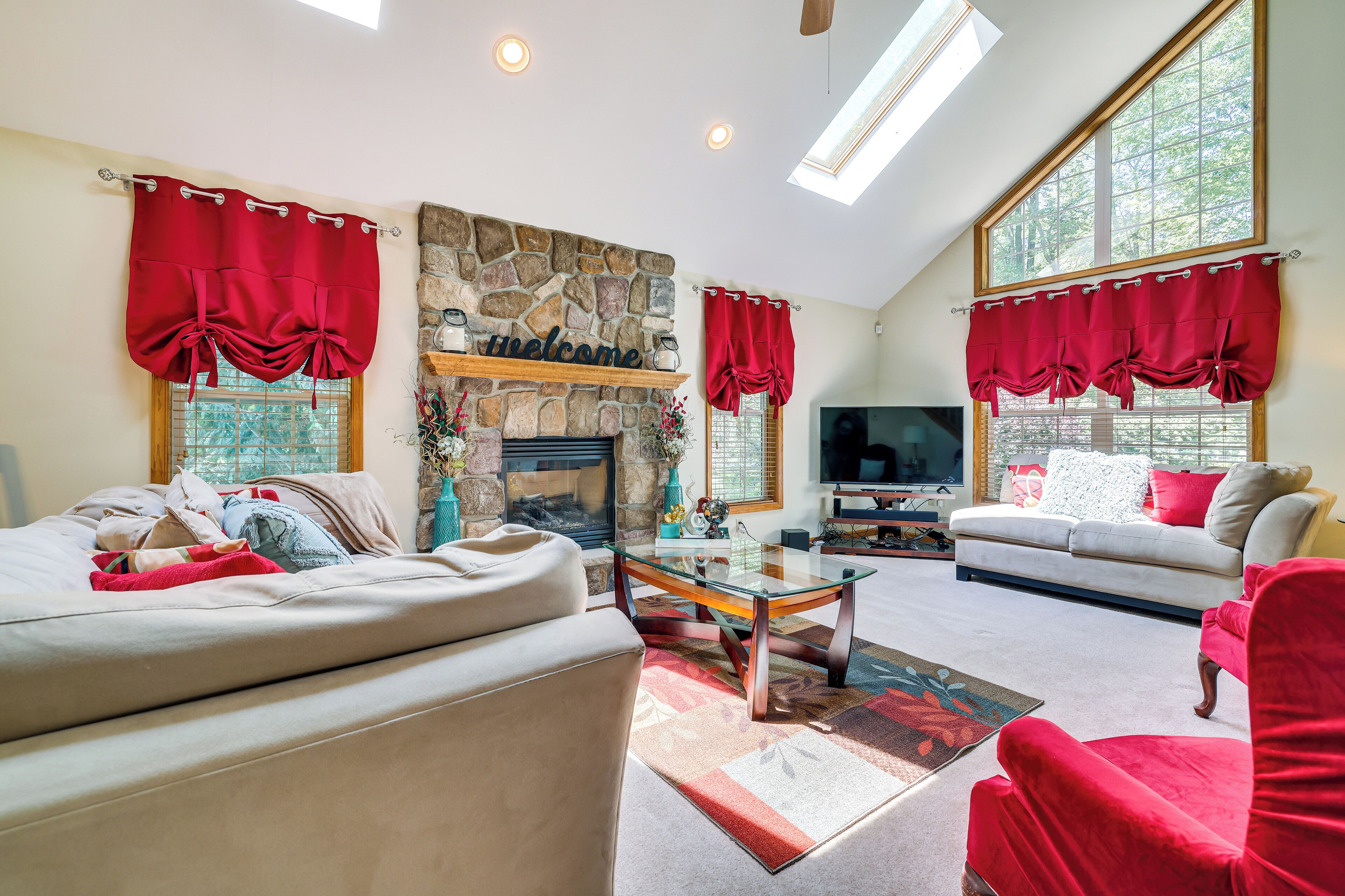 A living room with a stone fireplace, a sofa, a red chair, a television, and a large window with red curtains.