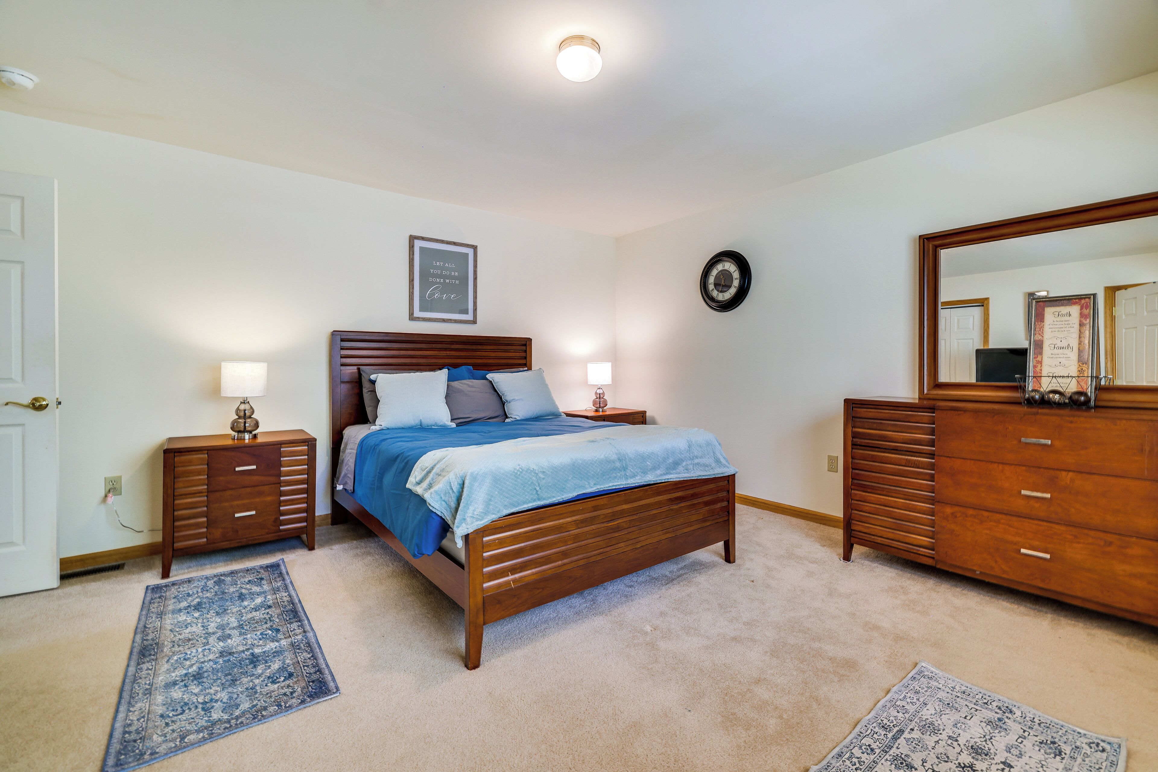 A bedroom with a wooden bed, two bedside tables, a dresser, a rug, and a wall clock.