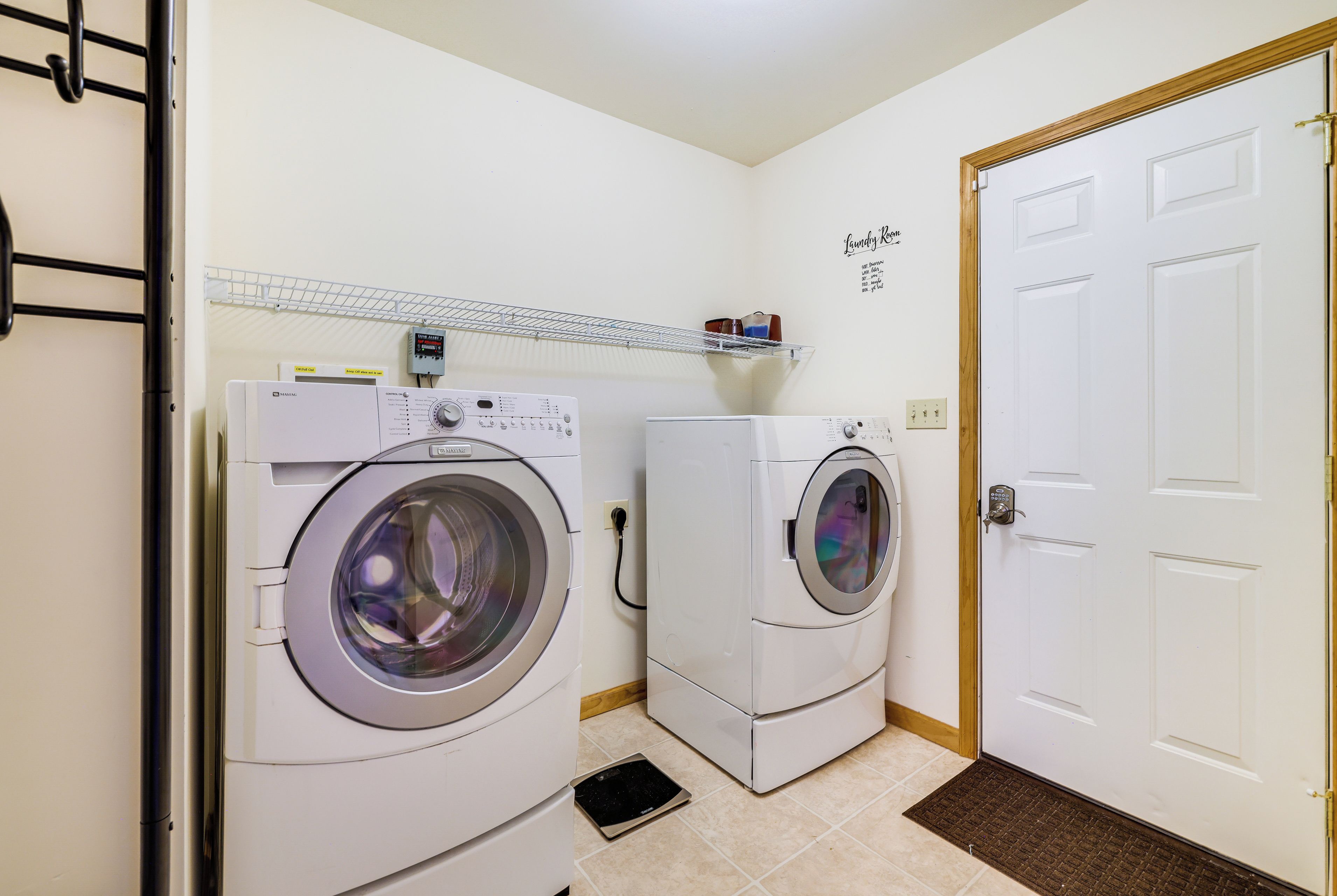 Un lavadero con dos lavadoras de carga frontal, un estante con productos de limpieza y una puerta con el letrero "Laundry Room".